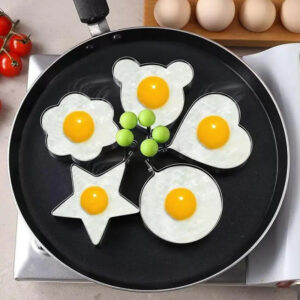 Five different shaped stainless steel egg fryers are frying eggs in a pan, topped with green grapes, and accompanied by eggs and cherry tomatoes.